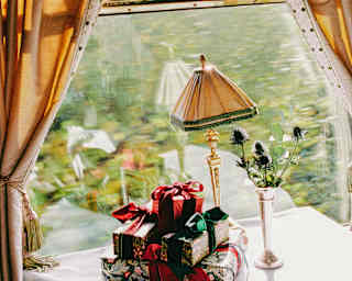 A pile of presents, wrapped with shiny paper and green and red bows rests on a window table below a festively decked pelmet.
