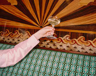 An arm in a pink satin sleeve raises a glass of Champagne up against the stunning marquetry panel behind a green banquette.