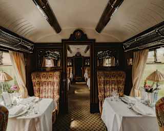 View of the Gwen Car with high-back chairs in beige and red-patterned fabric facing each other across tables laid for two.