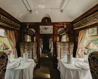 Seats with rich beige, blue and pink-patterned fabric flank tables laid for two with Champagne coolers, in the Ione Car.