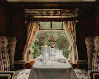 Looking at a table set for two and flanked by luxurious chairs, illuminated by the window and framed by tied-back curtains.