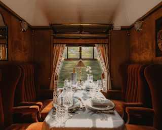 A window table with white cloth, crystal water and wineglasses, crockery, cutlery and wine cooler, flanked by russet velvet seats.