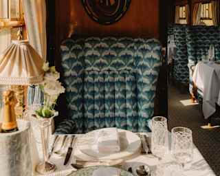 View across a table with crystal glasses, plates, cutlery, lamp, Champagne cooler and bud vase, to a luxurious teal chair.