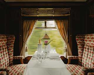 A view flies by a window table, laid for two, with Champagne and a bud vase, flanked by Zena's pink and grey-patterned seats.