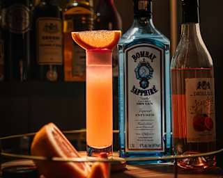 A blue Bombay Sapphire bottle sits between a luminous tall orange drink and a liquor bottle, both topped with citrus slices.
