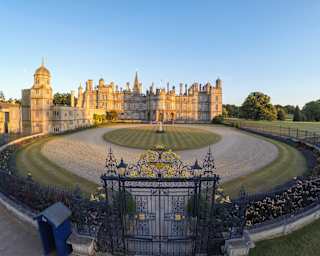 High-angle view of the large Burleigh House estate, stone glowing gold in late sun, seen over the wrought iron gates.