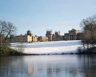 Blenheim Palace, in winter sun, sits atop a gentle sloping field covered with snow, which reflects on the icy lake water below.