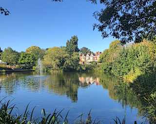 Bletchley Park stands grand beneath a clear blue sky, in a view across the lake that captures the tree-filled verdant estate.