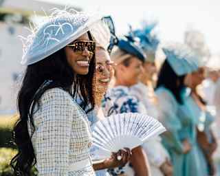 Dressed in hats and elegant summer frocks, a group of women smile and laugh as they enjoy a day at the races