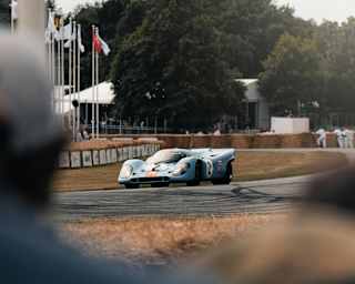 A light blue Porsche nine-one-seven race-car speeds on the track at Goodwood Festival of Speed, glimpsed through the crowd.