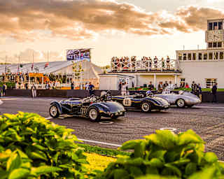 Saturated image of the track where three classic cars wait in line, and crowds mill at the side or watch from a roof terrace.