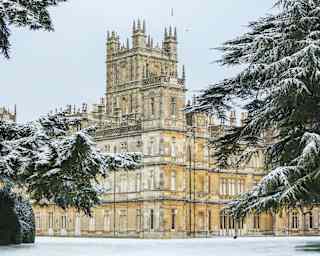 An impressive view of Highclere Castle with towering turrets and intricate roofscape, flanked by enormous snowy fir trees.