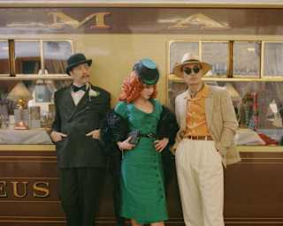 A woman with red curly hair in a green dress and hat leans against the stationed train flanked by two men, all in costume.