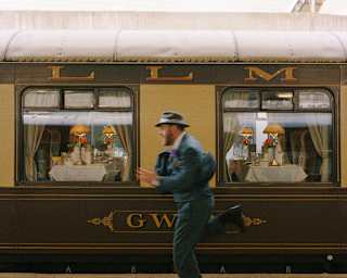 A man, dressed in a blue nineteen-thirties suit and hat runs in a blur on the platform in front of the stationed Gwen car.