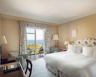 The Deluxe Room with large bed, neutral wall tones, floral soft furnishings and glorious sea view through balcony doors.