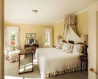 A Deluxe Suite double bed with fresh white bedding and elegant Jardine-pattern drapes with matching cushions and valance.
