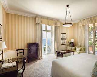Two balconies light up a bedroom with an armchair, desk, moonstone pendant light and striped wallpaper in wheat tones.