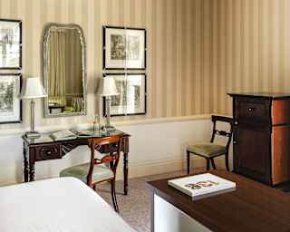 A bedroom corner with striped wheat wallpaper from ceiling to dado rail, with an antique wooden desk, chair and cabinet.