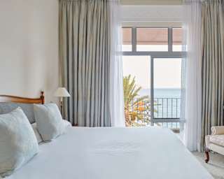 A large window flanked by drapes brings sublime sea views into a room with a white armchair and bed with two blue cushions.