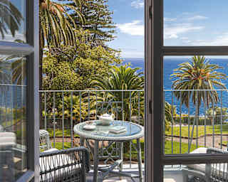Looking on to a suite balcony where a teapot and cup rest on a table with two chairs faced to enjoy garden and sea views.