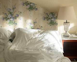 Soft morning light glances off the white duvet and pillow of an unmade bed with floral headboard and wooden side table.