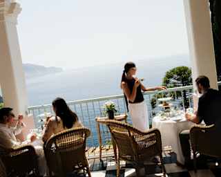 Tilted image of two couples, one woman standing, as they take afternoon tea on the ocean-view, monochrome-tiled tea terrace.
