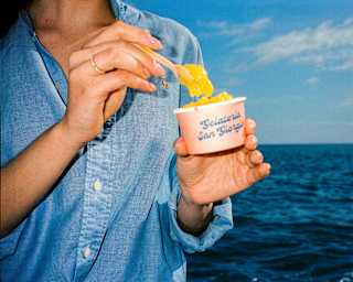 A woman in a blue shirt, seen from the neck down, scoops mango-hued gelato from a Gelateria San Giorgio pot, by the ocean.