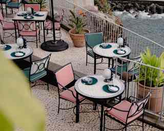 White-tiled tables with teal and pink chairs overlook rocks and sea through Brisa do Mar's terrace rails, seen from above.
