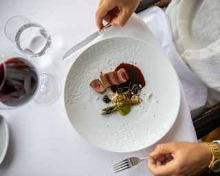 A diner holds a knife and fork by a plate of seared pork and asparagus at a table with red wine and water, seen from above.