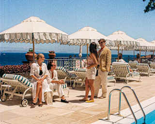 A couple, standing, talk with a man and woman seated on a striped lounger beneath a fringed white parasol at the poolside.