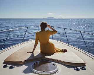 Lady in a yellow dress sitting on the bow of a sailing boat heading towards islands