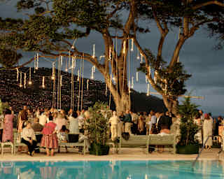 A group of guests gather around seating beneath a tree filled with dangling lights which reflect in the pool like fireworks.