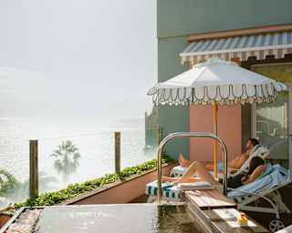 A man and woman lie on lounge chairs shaded by a parasol on a balcony with dramatic ocean views, seen over a water feature.