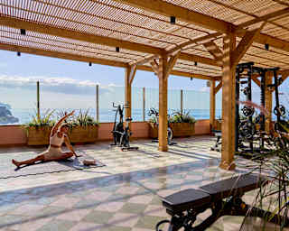 A woman in white yoga wear stretches an arm over her head as she sits, one leg extended, on the covered wellness terrace.