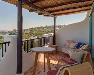 Two cushioned wicker chairs and a sofa enjoy coast views on a tiled terrace with sculpted walls, railings and bamboo shade.