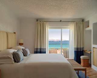 Glazed balcony doors flanked by cream and blue drapes bring azure sea views into a room with a bed dressed in white linen.