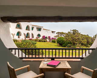 A table with a drink, book and two chairs sits in an enclosed terrace with a view of gardens through an inverted arch window.