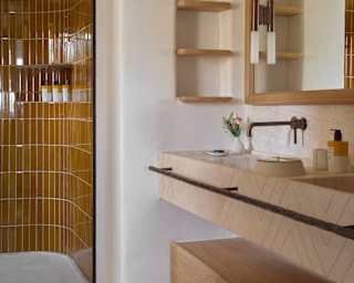A stone basin runs beneath a wooden mirror, in a bathroom view capturing a curving shower cubicle lined with amber tiles.