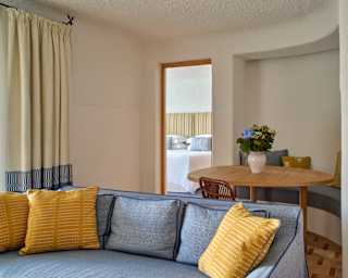 A table with a vase of flowers sits by an alcove with a built-in banquette, seen over a blue sofa with three yellow cushions.