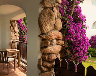Violet bougainvillea climbs support walls, created with stone and rendering, in a view of a balcony with table and chairs.