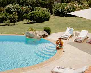 A man reads at the poolside of a villa pool with one foot in the water, seen in a high-angle view that captures the coast.