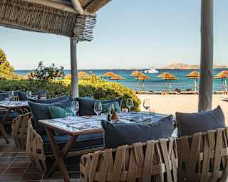 Latticed wicker cocoon chairs with blue cushions provide luxurious casual seating at a Entù e Mari table by the sunny beach.