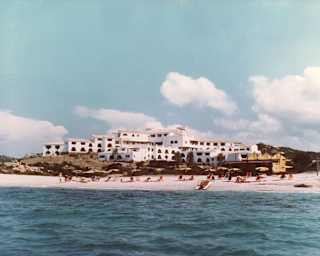 Historic image of beach-goers on the sand in front of Romazzino's hillside cluster of white buildings, seen from the sea.