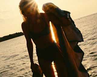 A woman, with a magazine in one hand and towel in the other, stands in white-water shallows, silhouetted against golden sun.