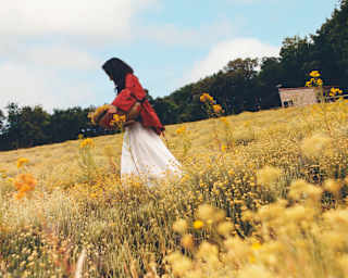 Low-angle image of a dark-haired woman in a white skirt and red top as she carries a basket of harvested flowers in a field.