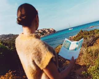 In soft-focus foreground, seen from behind, a woman holds a hiking guide and gazes at an azure bay along the Pevero Trail.