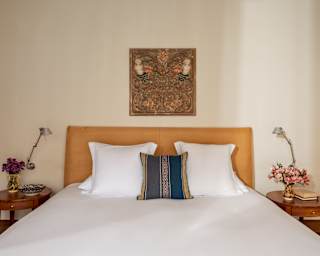 View facing a white double bed with a wood-carving on the wall above, flanked by side tables with flowers and reading lamps.