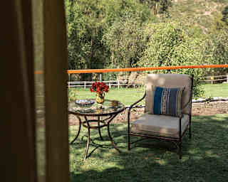 A patio chair and table rest in dappled shade on a lawn area with a breathtaking hillside backdrop, seen from inside a room.
