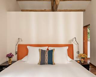 Facing a bed, with crisp white sheets, navy and gold woven cushion and orange headboard, flanked by side tables and lamps.