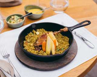 Close-up of a black griddle pan containing a confit duck leg on a bed of rice, served with yellow corn and carrots.